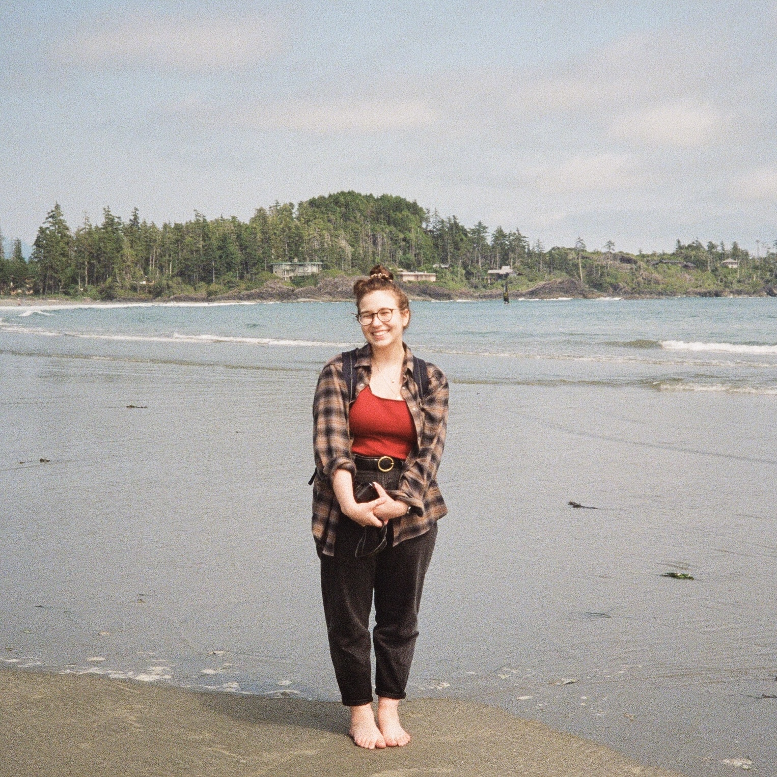 Smiling woman with brown hair, wearing flannel, standing on beach with trees in background.